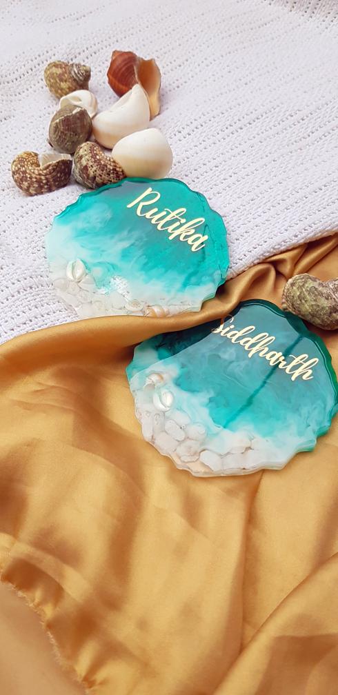 Two turquoise coasters with names engraved on them, surrounded by seashells on a textured surface.
