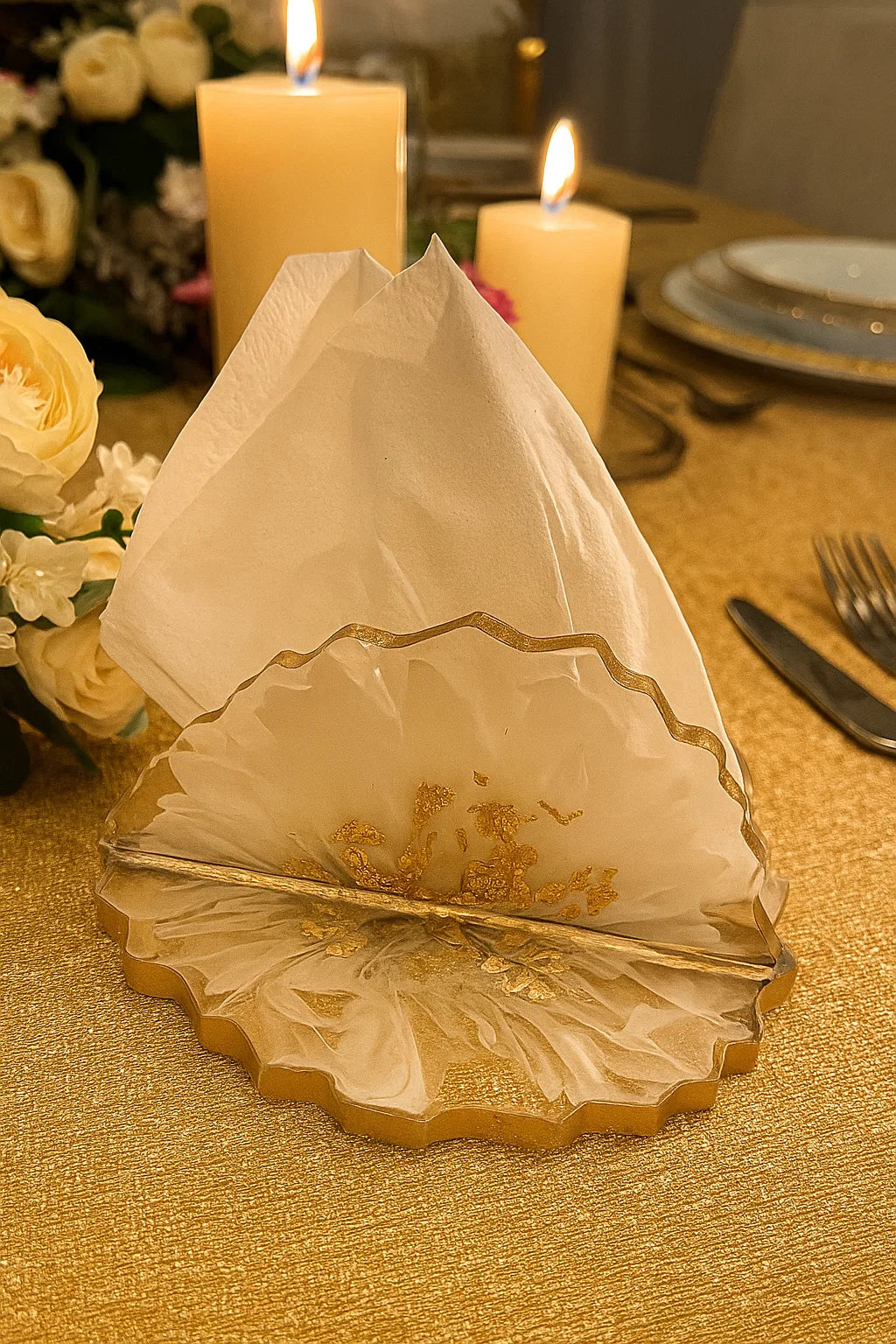 Decorative white napkin with gold trim on a gold tablecloth, candles, and flowers in the background.