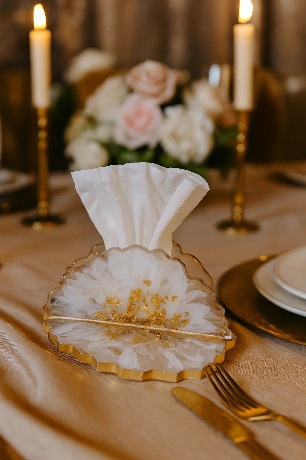 Decorative glass holder with a folded white napkin on a table setting with candles and flowers.