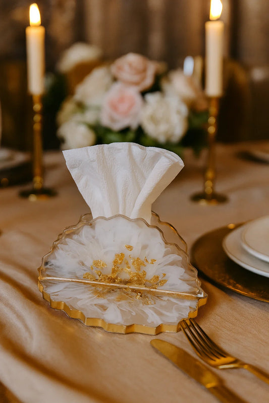 Decorative glass holder with a folded white napkin on a table setting with candles and flowers.